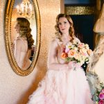 bride with hollywood hair wears a puffy pink gown and stands in front of a mirror on her wedding day