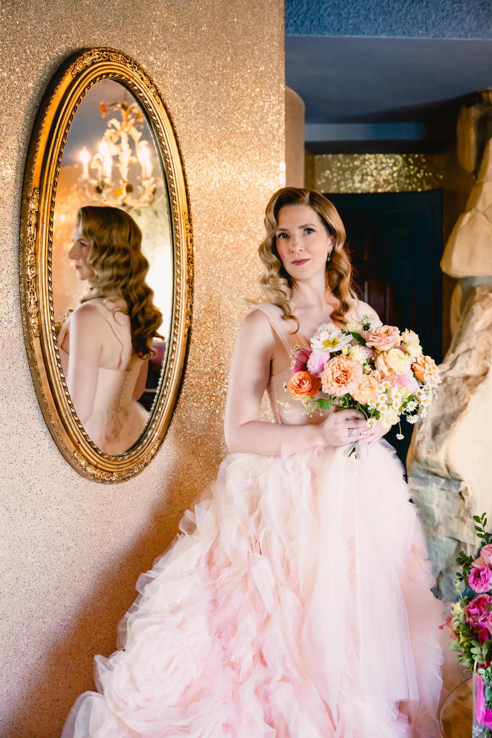 bride with hollywood hair wears a puffy pink gown and stands in front of a mirror on her wedding day