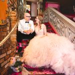 a couple sits on the staircase at madonna inn. the bride is wearing a pink wedding dress and the groom is wearing a white tuxedo. the carpet is pink.