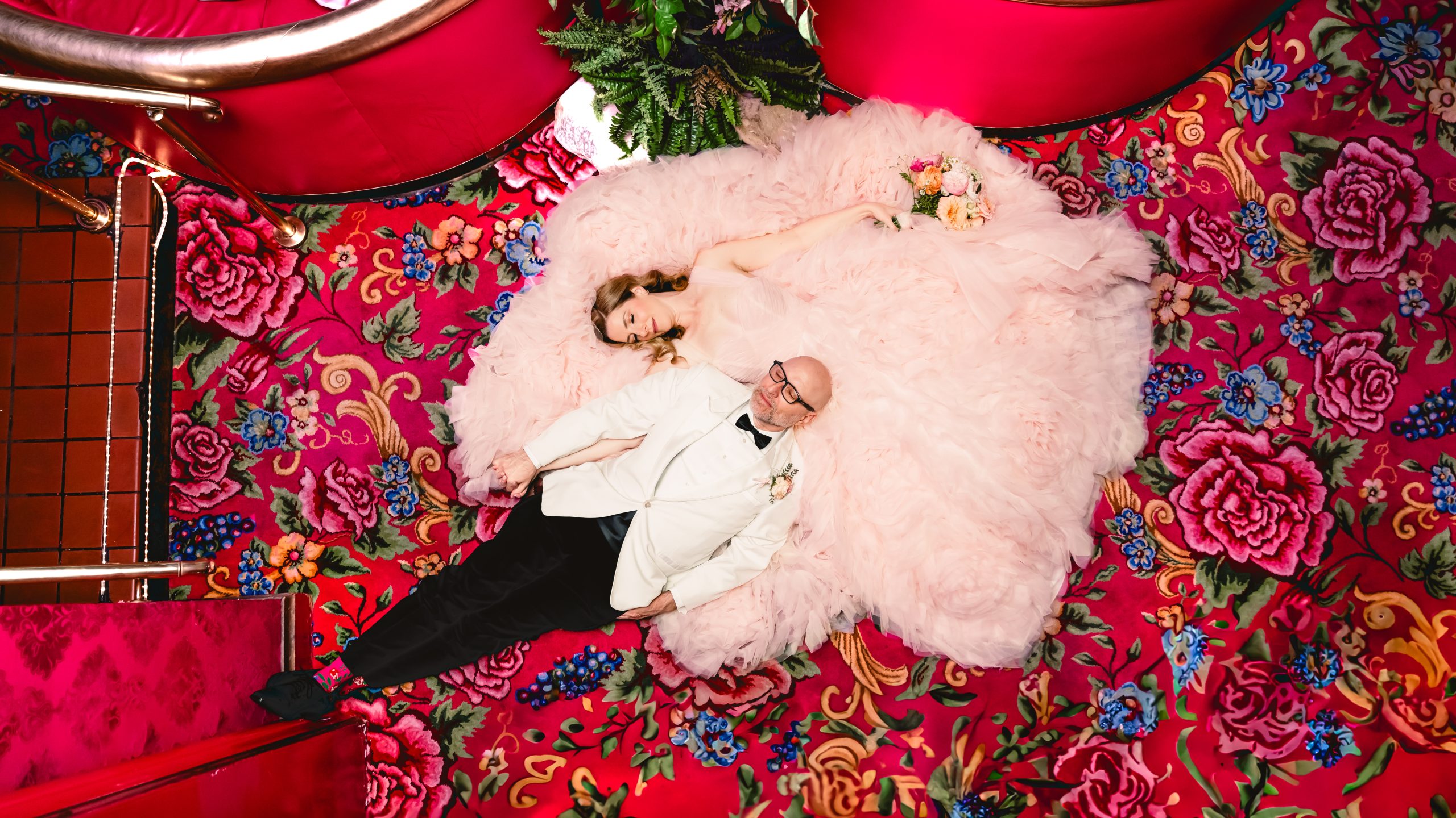 this is an aerial view of a couple laying on the pink floral carpet at madonna inn. the bride is wearing a fluffy pink dress and the groom is in a white tuxedo.