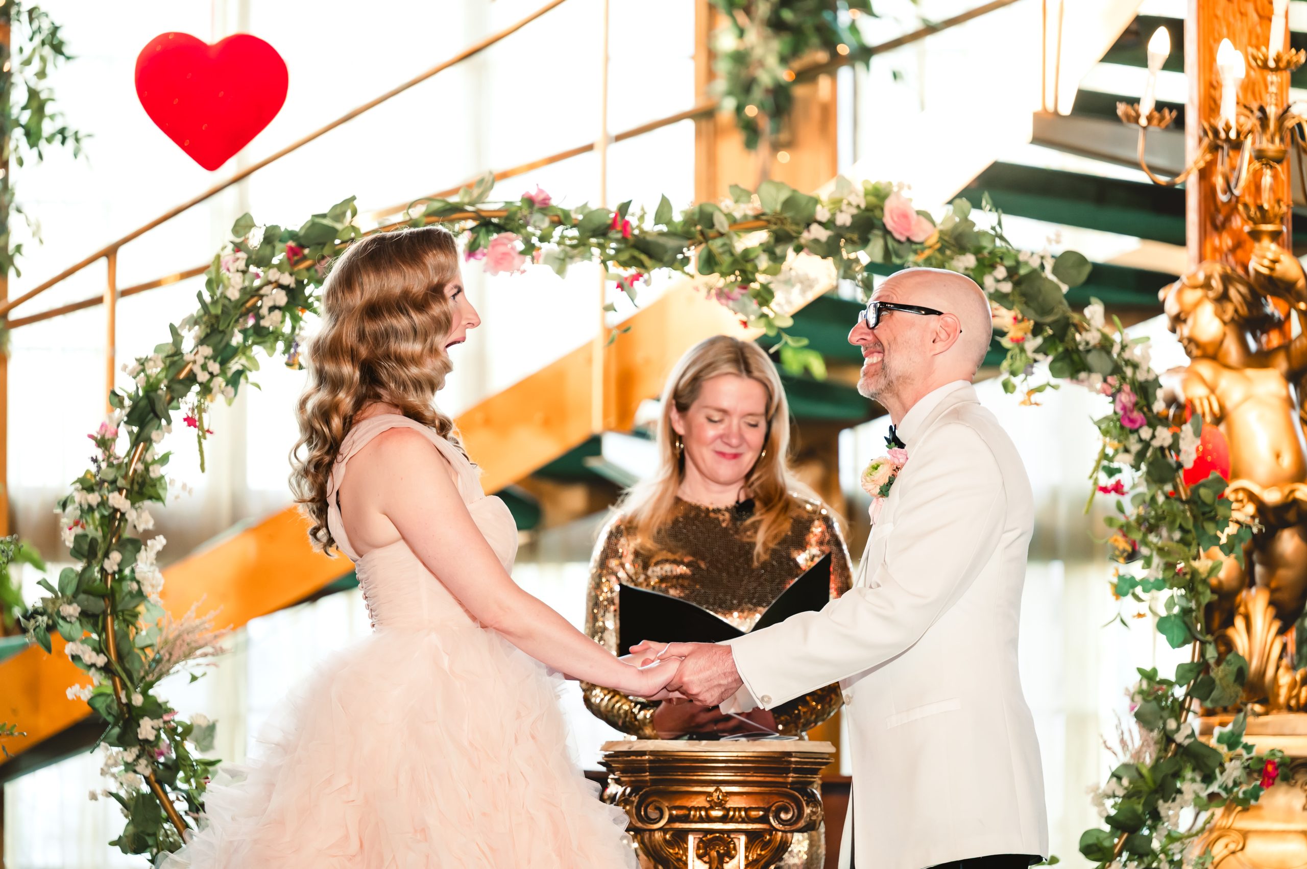 the bride and group gasp and laugh together as they hold hands under their floral arch