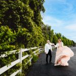 a groom in a white tuxedo and a bride in a pink wedding dress dance around together among green southern california tress with a white fence