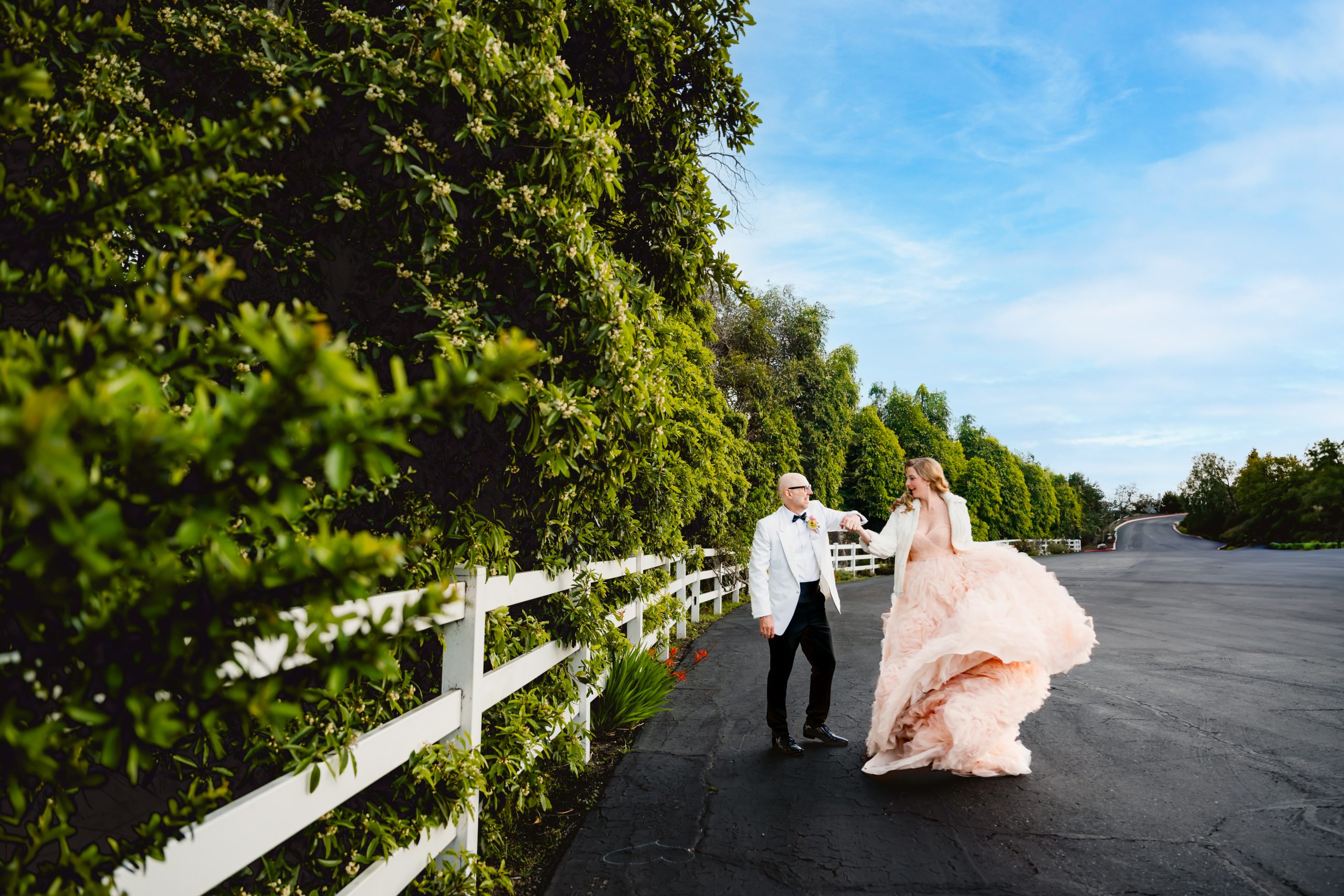a groom in a white tuxedo and a bride in a pink wedding dress dance around together among green southern california tress with a white fence