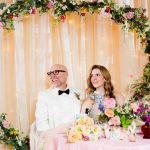a couple sits together under a heart shaped arch covered in flowers. their table is full of maximalist details with floral and vintage elements.