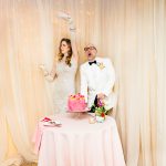 a bride in a vintage champagne mini dress and a groom in a white tuxedo sing about their pink champagne cake as they cut it
