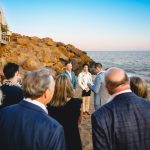 two grooms surrounded by their guests during their wedding on a malibu beach. you can see the cliffs and ocean in the background.