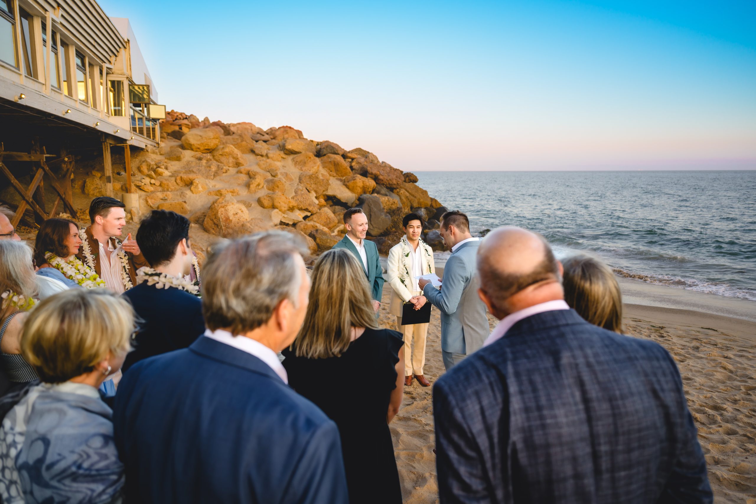 two grooms surrounded by their guests during their wedding on a malibu beach. you can see the cliffs and ocean in the background.