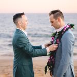 a groom in green places a purple lei around his new husband's neck during their wedding ceremony as the sun sets over the ocean in malibu