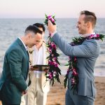 a groom in a light blue suit places a purple lei around his new husband's neck during their wedding ceremony as the sun sets over the ocean in malibu