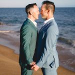 a groom in a green suit and a groom in a light blue suit hold hands and put their noses together on the beach