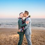 A couple embraces and have their first kiss as a married couple during their elopement ceremony on a malibu beach. The sun is setting over the ocean behind them.