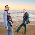 a groom in a light blue suit laughs as his husband, wearing green, kicks sand and laughs behind him.