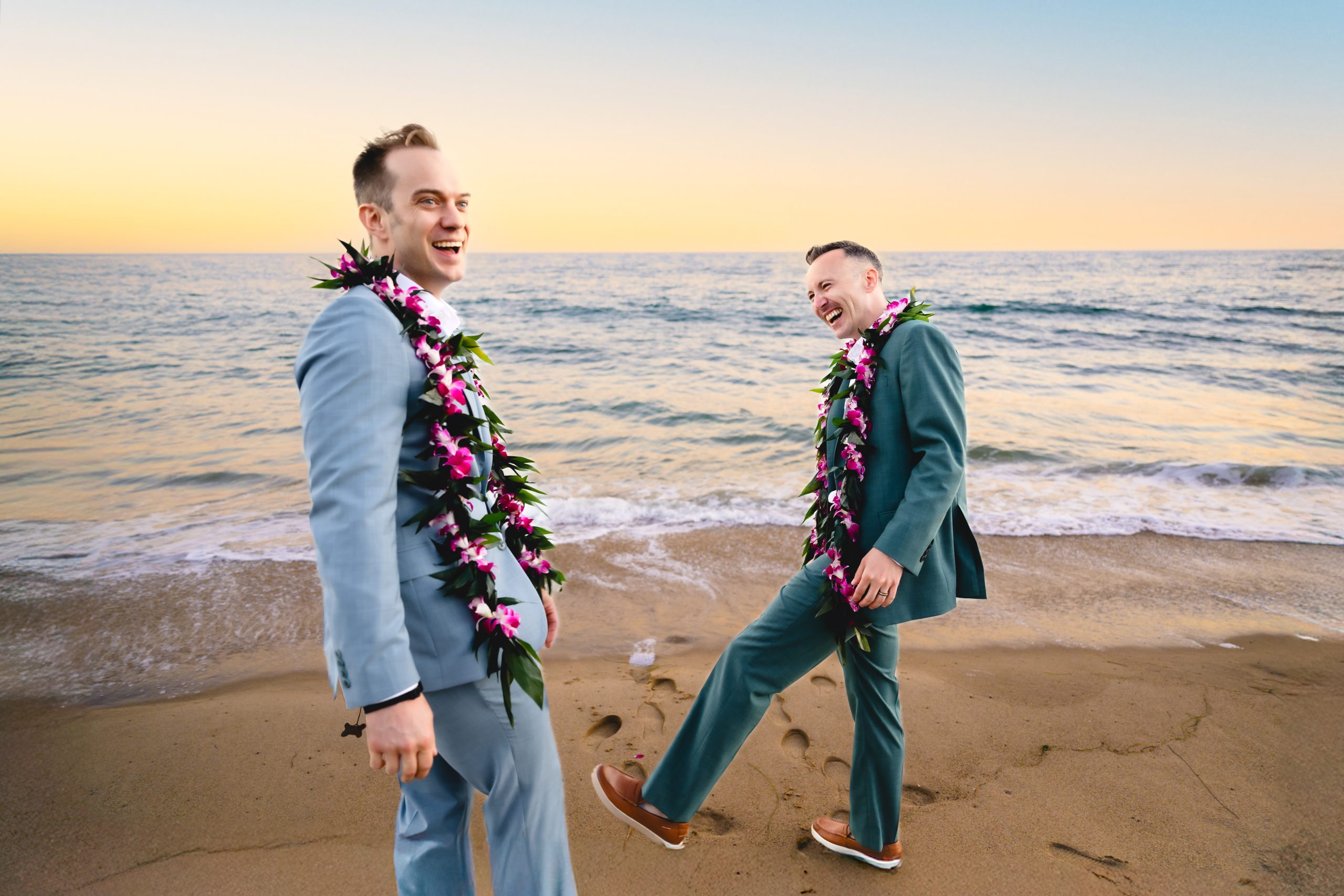 a groom in a light blue suit laughs as his husband, wearing green,  kicks sand and laughs behind him. 