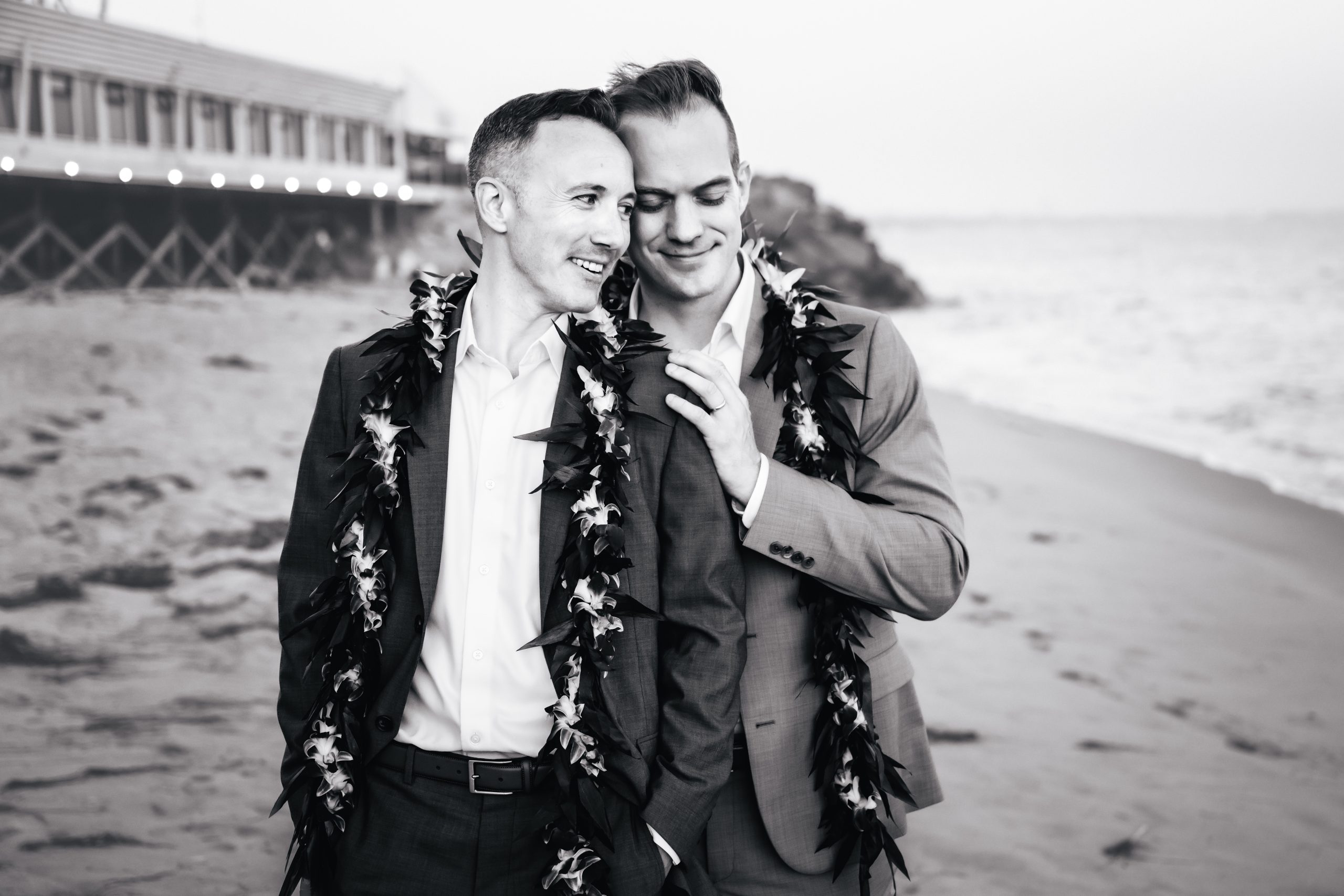 a couple embraces, one groom behind the other, holding each other tight with Mastro's Ocean Club behind them.