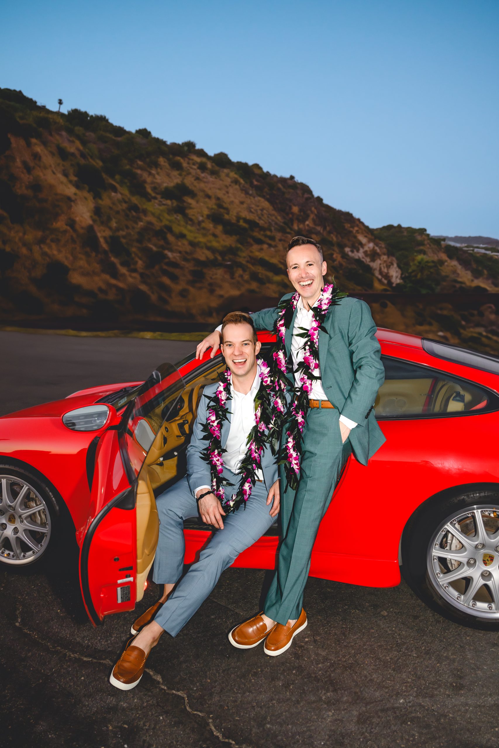 Two grooms pose in front of their red porsche on their wedding day