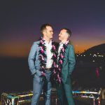 A groom in green and a groom in blue hold each other as the sun fades and night begins