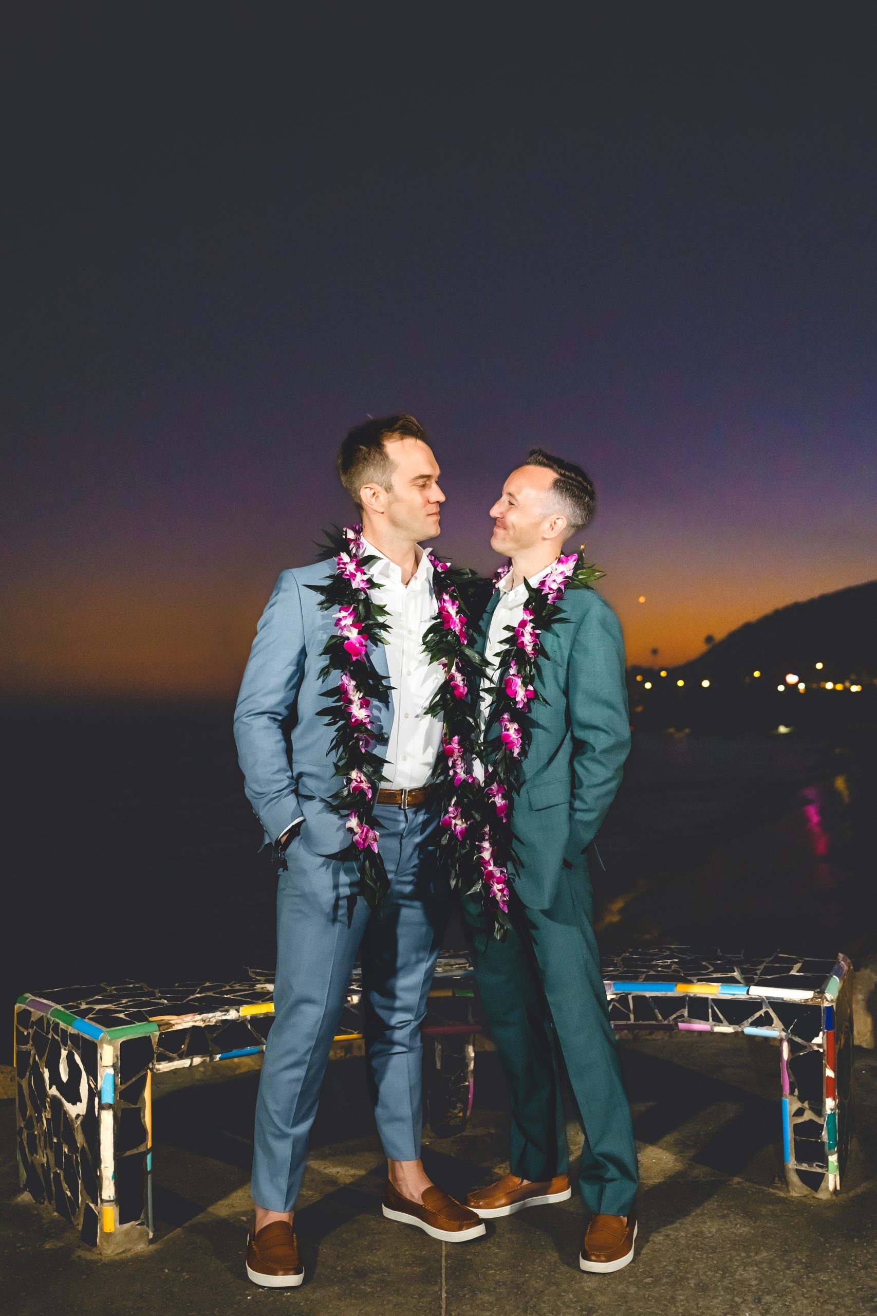 A groom in green and a groom in blue hold each other as the sun fades  and night begins
